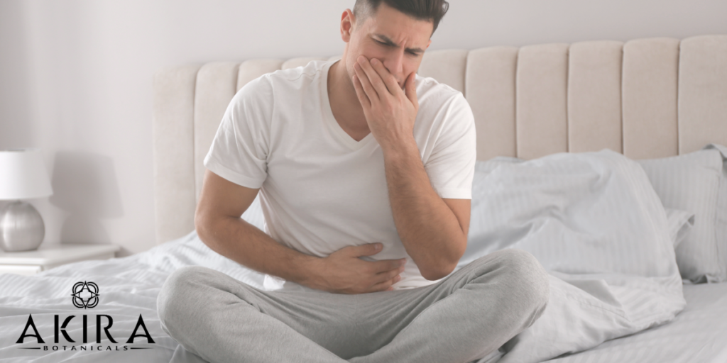 A man sitting on a bed in a white undershirt and sweat pants holding his stomach and mouth.
